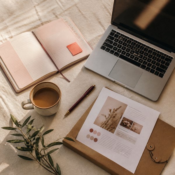 Overhead flat lay of a laptop, a blush planner, a cup of coffee and a website design tucked into a kraft folder on cream linen